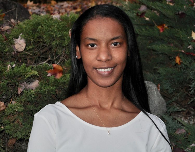 Quin'Nita Cobbins-Modica stands in front of a flowering bush and smiles.