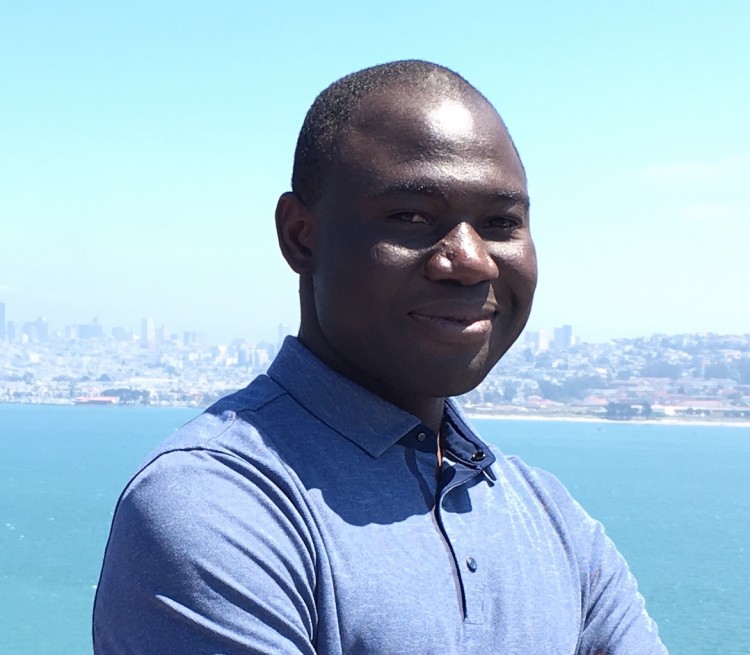 Matthew Adeiza wears a blue shirt in front of a bright blue ocean scene.
